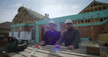 İnşaat alanında duran inşaatçılar, çerçeve altındaki ev, takım çalışması ve işbirliği, ahşap şapkalı iki adam, aletler ve malzemelerle çalışabilir, tamamlanmamış bir ev.