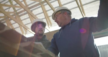 İnşaat danışmanı ve iş arkadaşı proje detaylarını, takım çalışmasını ve rehberliği tartışıyorlar. Her ikisi de arka planda yapısal çerçeveye sahip güvenlik miğferlerinde.