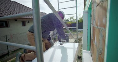 Yalıtım tahtasına yapıştırıcı süren inşaat işçisi, duvar kurulumu, güvenlik kaskı, arka planda merdiven ve yapı iskelesi olan dış yapı projesi için hazırlık yapıyor.