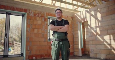 Kendine güveni tam bir inşaat işçisi, kolları çapraz, sıkı bir şapka ve tulum giyiyor, bitmemiş bir binanın içinde duruyor, kararlılık ve profesyonellik sergiliyor.