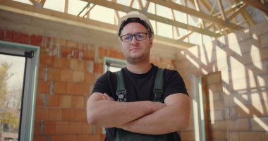 Kendine güveni tam bir inşaat işçisi, kolları çapraz, sıkı bir şapka ve tulum giyiyor, bitmemiş bir binanın içinde duruyor, kararlılık ve profesyonellik sergiliyor.