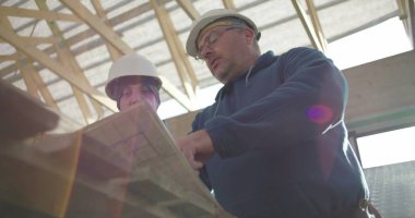 İnşaat danışmanı ve iş arkadaşı proje detaylarını, takım çalışmasını ve rehberliği tartışıyorlar. Her ikisi de arka planda yapısal çerçeveye sahip güvenlik miğferlerinde.