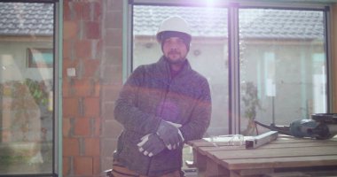 Kendine güvenen inşaat işçisi, kısmen inşa edilmiş bir yapının çalışma tezgahının yanında elleri ve kalçaları üzerinde poz veriyor, güvenlik donanımı, takım çalışması ve inşaat alanında gurur takıyor.