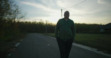 Gün batımında boş bir kırsal yolda yürüyen bir adam, elleri ceplerinde, huzurlu bir kırsal bölgede sonbahar ağaçları ve açık gökyüzü ile huzurlu bir akşamın tadını çıkarıyor.