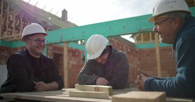 İnşaat işçileri çalışma masasının etrafında birlikte gülüyorlar, başlık takıyorlar, neşeli bir anı paylaşıyorlar, ev inşaatı sırasında bağ kuruyorlar.