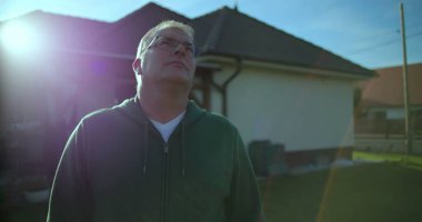 Dışarıdan bakan bir adam, arka planda bir ev, yeşil çimenler, huzurlu bir banliyö mahallesi, düşünceli bir ifade.