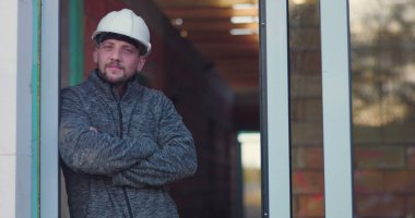 İnşaat işçisi kapıda kolları çaprazlanmış, düşünceli bir şekilde dik dik bakıyor, başlık takıyor, arka plan yapım aşamasında olduğunu gösteriyor, kararlılığı ve odaklanmayı simgeliyor.