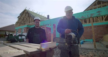 Ahşap blokta elektrikli testereyle çalışan inşaat işçisi, meslektaş gözlemi, inşaat alanında takım çalışması, arka planda yapı inşaatı, işçiliğe odaklan