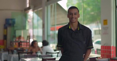 Neşeli vücut diline sahip sıradan bir restoranın penceresinin yanında gülümseyen erkek garson, arka planda oturan müşteriler, kentsel gün ışığı.