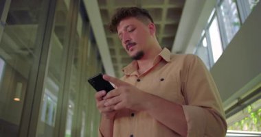 Ofis koridorunda akıllı telefon kullanan genç adam, sakin ve odaklanmış bir ifadeyle ekrana bakıyor, gündelik ve modern çalışma ortamı