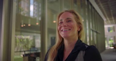 Modern koridorda neşeli bir şekilde gülen kadın, gerçek mutluluğu ve açık ifadeyi gösteriyor, pozitif işyeri duygusunu ve insan bağlantısını temsil ediyor.