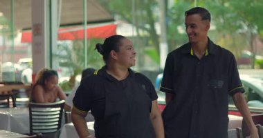 Latin garson ve genç garson restoran yemek alanında birlikte gülümsüyor, arkadaş canlısı ve misafirperver bir tavırla yan yana duruyorlar.