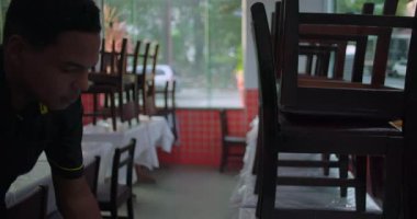 İki restoran çalışanı vardiyanın başında masalardan sandalye kaldırıyor. Günlük yemek alanlarında gün ışığıyla başlıyorlar.