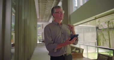 Orta yaşlı bir adam ofis koridorunda gülümserken cep telefonu gevşetici ifadeler kullanıyor. Parlak, modern iş yerinde mobil cihazlarla meşgul oluyor.