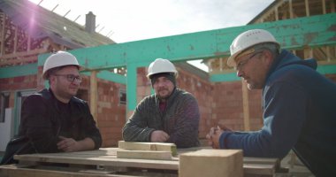 İnşaat işçileri çalışma masasının etrafında birlikte gülüyorlar, başlık takıyorlar, neşeli bir anı paylaşıyorlar, ev inşaatı sırasında bağ kuruyorlar.