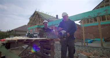 Ahşap blokta elektrikli testereyle çalışan inşaat işçisi, meslektaş gözlemi, inşaat alanında takım çalışması, arka planda yapı inşaatı, işçiliğe odaklan