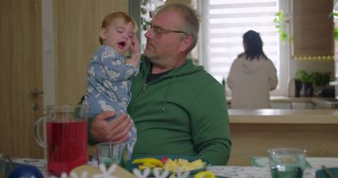 Baba kucağında bir bebekle masada oturuyor, çocuk esniyor, aile kahvaltısı hazırlıyor, sıcak ve sakin bir atmosfer yaratıyor, mutfak tezgahında arka planda bir kadın oturuyor.