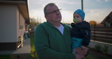 Baba, bebeğini emzik ile arka bahçede tutuyor. Çocuk kıyafetlerini nazikçe ayarlıyor. Huzurlu bir aile ortamında sevgi ve sevgi gösterisi yapıyor..