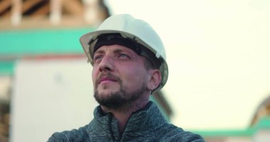 Başlıklı inşaat işçisi kendine güvenerek yukarı bakıyor, arka planda açık yapı, görüşü, gururu ve inşaat mükemmelliğine bağlılığı simgeliyor.