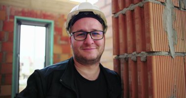 Tuğla duvara dayanmış inşaat işçisi, gülümsüyor ve güvenlik kaskı ve gözlüğü takıyor, tamamlanmamış bir binanın içinde, profesyonel varlığını ve güvenini gösteriyor.