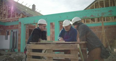 Planlar etrafında tartışan inşaat işçileri, sert şapkalar takanlar, konut inşaatının planlarını gözden geçirenler, arka planda görünen çerçeve ve duvarcılık