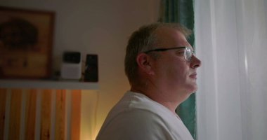 Pencereden dikkatlice bakan adam, yan görünüm, gözlük takan, sakin yüz ifadesi, içeriyi yansıtan, sakin bir ev ortamında sakin bir iç gözlem anı.