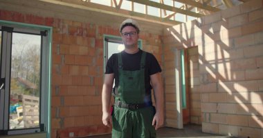 İnşaat işçisi kısmen inşa edilmiş bir yapının içinde duruyor, tulum ve başlık giyiyor, odaklanmış ve kendini işine adamış görünüyor, şantiyede profesyonelliği somutlaştırıyor.