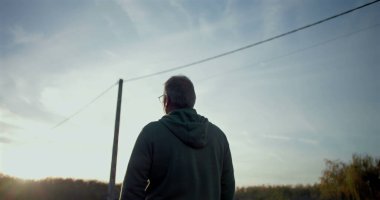 Kırsal bir yolda yürüyen bir adam, düşünceli bir şekilde ileriye bakıyor, sakin, açık bir çevre, yol boyunca sıralanmış elektrik direkleri ve ağaçlar, sakin kırsal atmosfer.