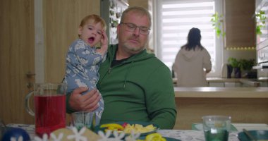 Baba kucağında bir bebekle masada oturuyor, çocuk esniyor, aile kahvaltısı hazırlıyor, sıcak ve sakin bir atmosfer yaratıyor, mutfak tezgahında arka planda bir kadın oturuyor.