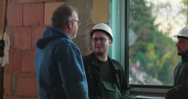 Cam kenarında sohbet eden inşaat işçileri, rahat ve günlük etkileşimler, kapalı alanda yenileme alanı, dostluk ve takım çalışması, güvenlik teçhizatı ve sert şapkalar.