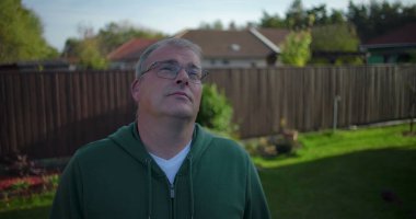 Arka bahçeye bakan bir adam ahşap çitlerin ve konut evlerinin yakınına günlük ve düşünceli bir an, banliyö mahallesi, yeşillik ve sakin bir çevre.