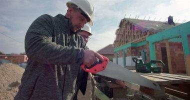 İnşaat işçisi ahşap tahtayı el testeresiyle kesiyor, iş arkadaşı gözlemliyor, inşaat sahasında takım çalışması, ev iskeleti görünür, hassasiyet ve işçiliğe odaklanmış.