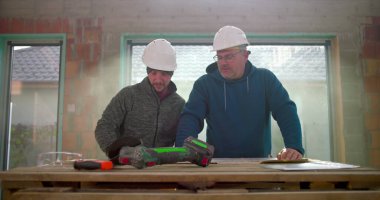 İnşaat işçileri masadaki planları inceliyor, etraftaki araçlarla yenileme projesinin ayrıntılarını inceliyor, inşaat alanındaki işbirlikçi yaklaşımlar