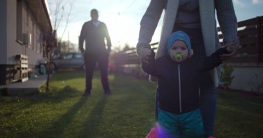 Bebek, anne desteğiyle arka bahçede yürümeyi öğrenirken, baba uzaktan izlerken el ele tutuşurken, dışarıda değerli aile anlarının tadını çıkarıyor..