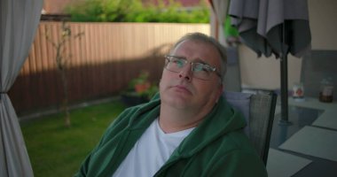 Bahçe sandalyesinde oturan bir adam, nazikçe gülümseyen ve düşünceli bir şekilde bakan, bahçedeki bitki ve yeşilliklerle dolu sessiz bir anın tadını çıkaran bir adam.