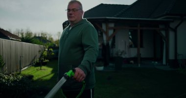 Bahçe hortumunu arka bahçede tutan adam, konut evinin yanında duran, yeşil, veranda ve çimenli, rahat açık hava aktivitesi olan banliyö ortamında sıradan bir an.