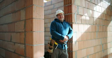 İnşaat işçisi mavi kapüşonlu ve kasklı inşaat halindeki tuğladan duvara yaslanmış, kemerli, kendine güvenen ve rahat duruşu olan.