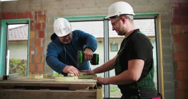 İnşaat işçileri inşaat sahasında ahşap döşeme, yapı iskelesinde takım çalışması, bir işçi sondajı, diğeri asistanlar, hassasiyet ve işbirliğine odaklanıyor.