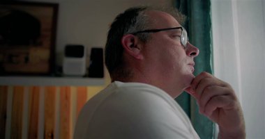 Pencere kenarında duran bir adam, çeneye yakın, derin düşüncelere dalmış, gözlük takmış, yansıtıcı ifade takmış, hayatı kapalı bir yerde düşünüyor, sakin bir an, içe kapanık ve sakin bir ruh hali.