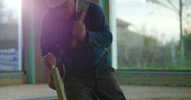 İnşaat işçisi odun parçalarını odaklama ve kuvvetle çekiçliyor, iç inşaat sahasında diz çöküyor, malzemeleri güvence altına alıyor, inşaat projesi devam ediyor.