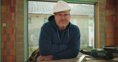 Çalışma tezgahının yanında poz veren inşaat işçisi, kısmen inşa edilmiş yapı, kendine güvenen ve ulaşılabilir ifade, inşaat alanı ortamı