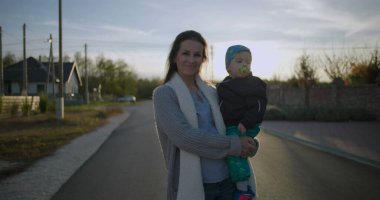 Anne, sessiz bir banliyö sokağında açık havada gülümserken kucağında bebekle kucağında bebekle güneşli bir günde mahallenin yakınlarındaki evlerin keyfini çıkarıyor.
