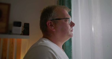 Pencereden dikkatlice bakan adam, yan görünüm, gözlük takan, sakin yüz ifadesi, içeriyi yansıtan, sakin bir ev ortamında sakin bir iç gözlem anı.