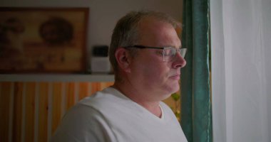 Pencereden dikkatlice bakan adam, yan görünüm, gözlük takan, sakin yüz ifadesi, içeriyi yansıtan, sakin bir ev ortamında sakin bir iç gözlem anı.