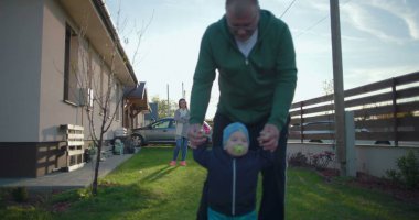 Anne uzaktan izlerken, baba bebeğin çimenlerde yürümesine yardım ediyor. Aile birlikte olmanın ve doğal ortamlarda öğrenmenin tadını çıkarıyor.