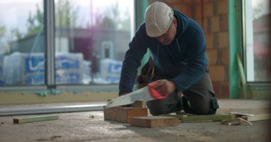 İnşaat işçisi inşaat alanında ahşap kesiyor, kesme işine odaklanmış, başlık takıyor, tuğla duvarlarla çevrili, inşaat projesi devam ediyor.