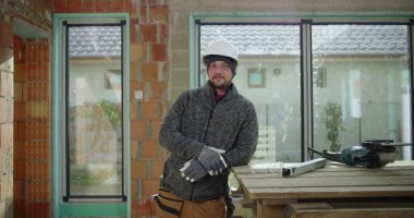 Kendine güvenen inşaat işçisi, kısmen inşa edilmiş bir yapının çalışma tezgahının yanında elleri ve kalçaları üzerinde poz veriyor, güvenlik donanımı, takım çalışması ve inşaat alanında gurur takıyor.