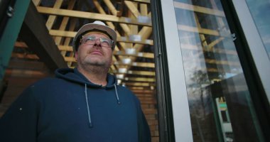 İnşaat işçisi açık çatı çerçevesine bakıyor, başlık ve gözlük takıyor, düşünceli bir ifadeyle bina inşaatının ilerlemesine odaklanıyor.