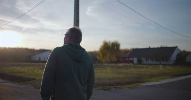 Sakin bir banliyö caddesinde yürüyen bir adam, sakin bir ifadeyle ileriye bakıyor, kırsal kesimde bir atmosfer, tuğla duvar ve arka planda evler, huzur ortamı.