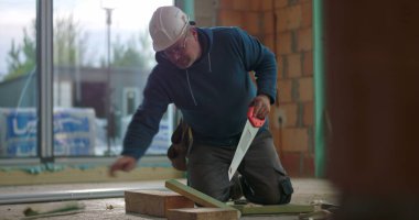 İnşaat işçisi inşaat alanında ahşap kesiyor, kesme işine odaklanmış, başlık takıyor, tuğla duvarlarla çevrili, inşaat projesi devam ediyor.